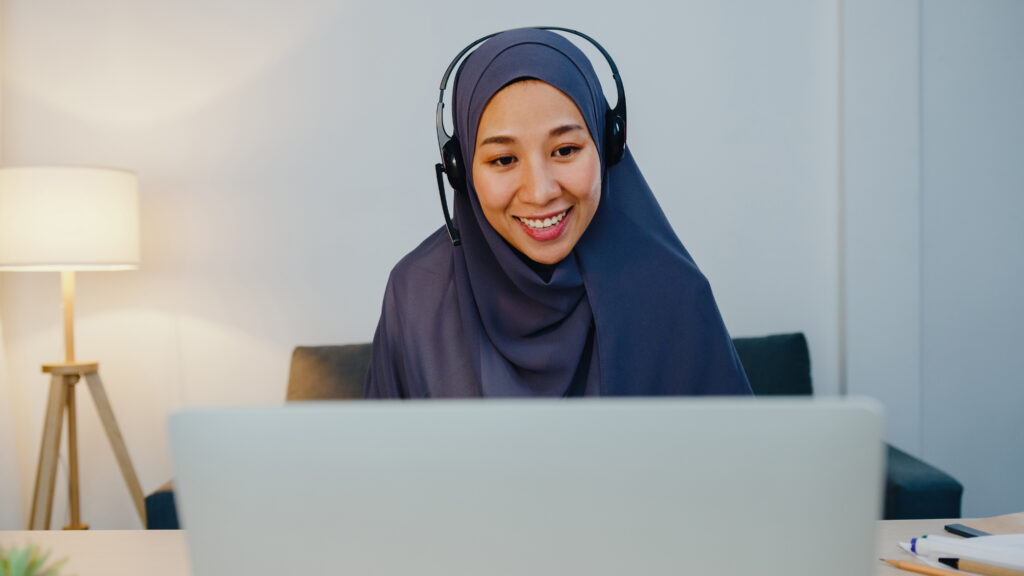 A smiling Muslim woman wearing a dark blue hijab and a headset with a microphone, looking attentively at her laptop screen. She is sitting at a desk in a well-lit, cozy home office or living room setup. In the blurred background, there is a modern wooden tripod floor lamp emitting warm light and a grey sofa, creating a professional yet welcoming online learning environment.